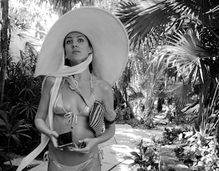 A lady in a swimsuit and oversized hat strolling down a garden path
